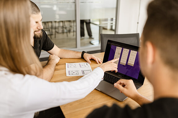 Ein UX Designerin und zwei Entwickler sitzen an einem Besprechungstisch und blicken gemeinsam auf den Bildschirm eines Laptops. Die Designerin zeigt auf ein Element auf dem Screen.