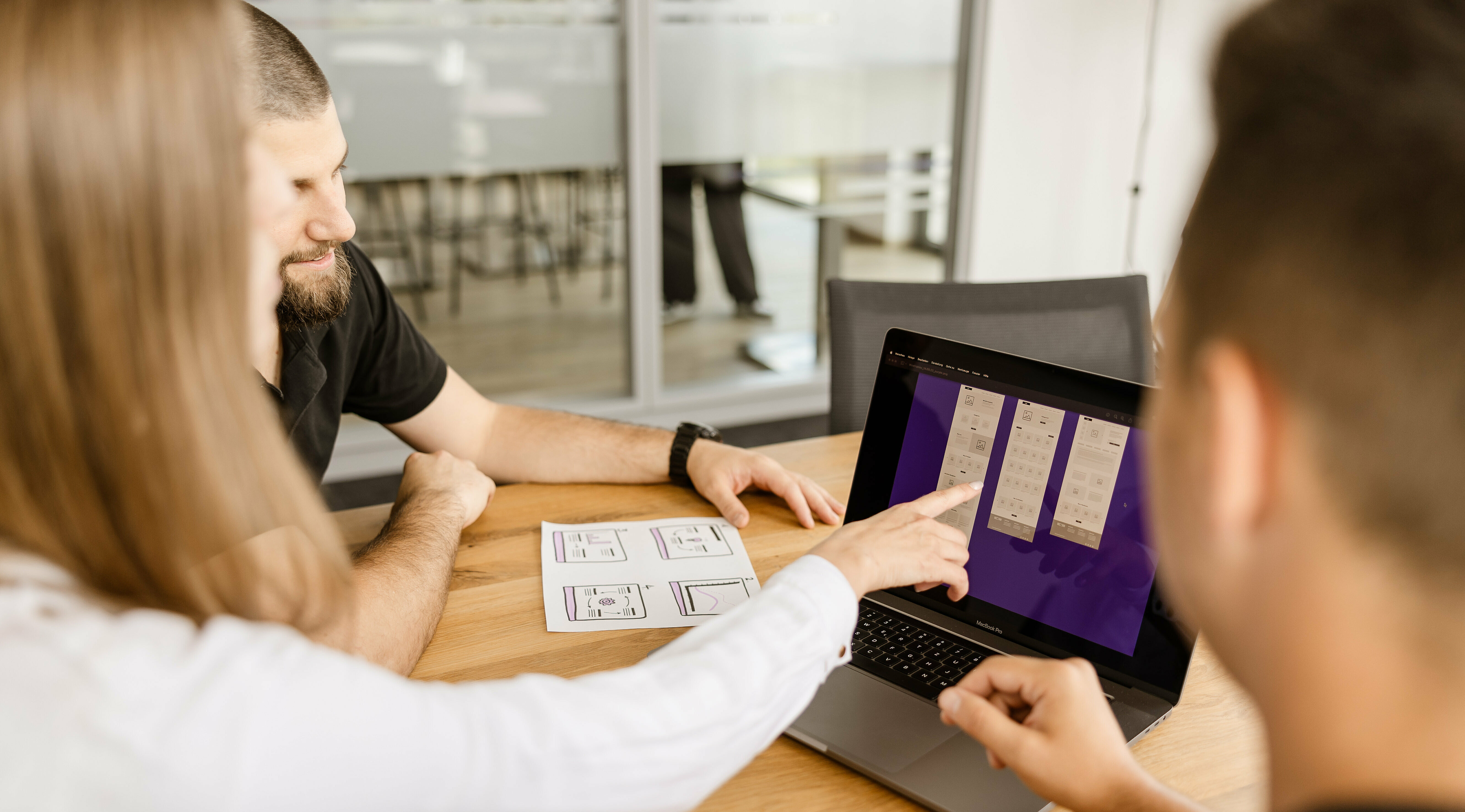 Ein UX Designerin und zwei Entwickler sitzen an einem Besprechungstisch und blicken gemeinsam auf den Bildschirm eines Laptops. Die Designerin zeigt auf ein Element auf dem Screen.